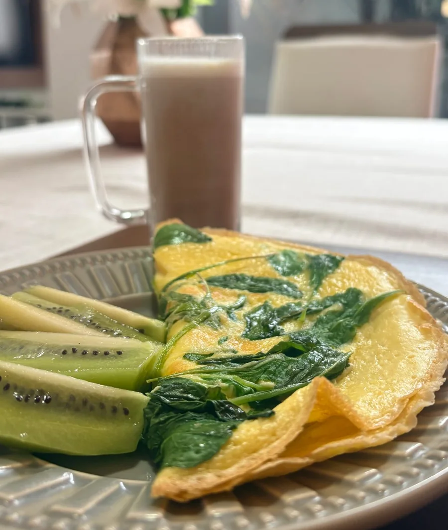 Prato com omelete de espinafre cozido e kiwi fatiado, ao lado de um copo de vitamina